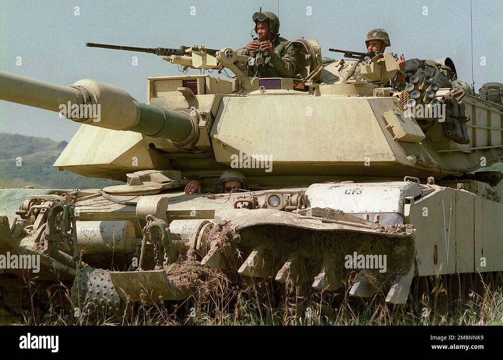 medium-shot-left-side-front-view-of-a-desert-camouflaged-m1a1-abrams-tank-with-a-mine-plough-system-the-tank-commander-loader-and-driver-are-outside-their-turrets-the-commander-mans-the-127mm-ma